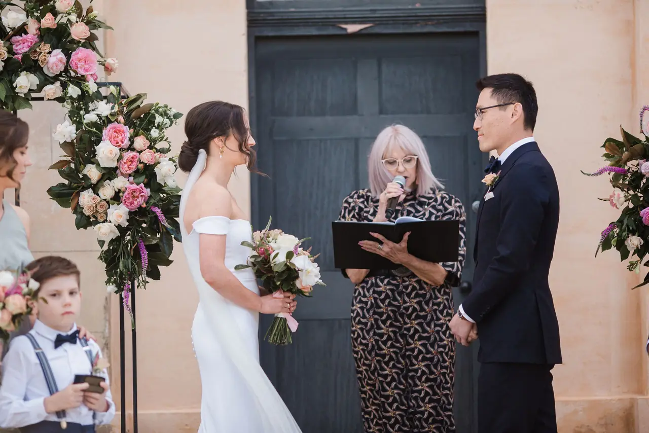 A celebrant leads a wedding ceremony beside a couple and a floral arrangement.