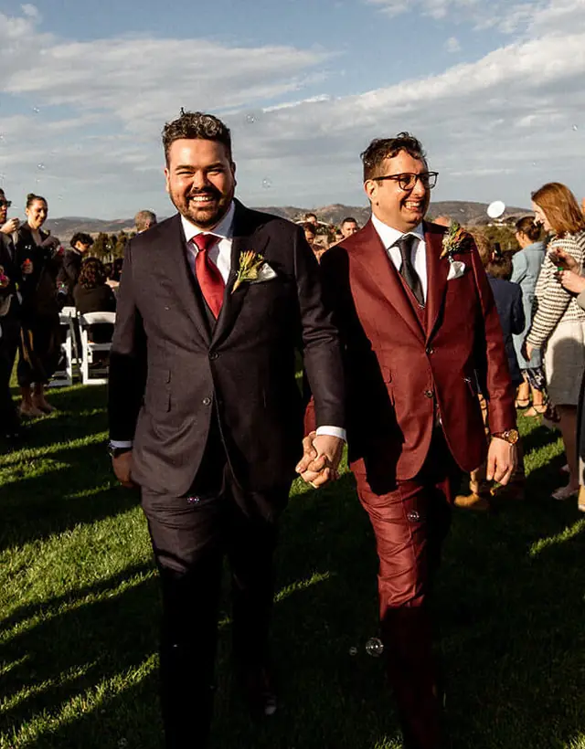 Two newly married men walk hand in hand through their outdoor wedding guests.